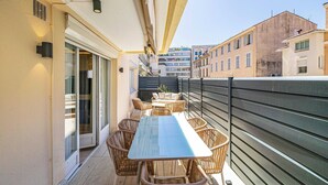 A balcony with wicker furniture, a glass table, and a view of adjacent buildings. - Luxurious Apartment - Croisette/hotel Mondrian (Cannes)