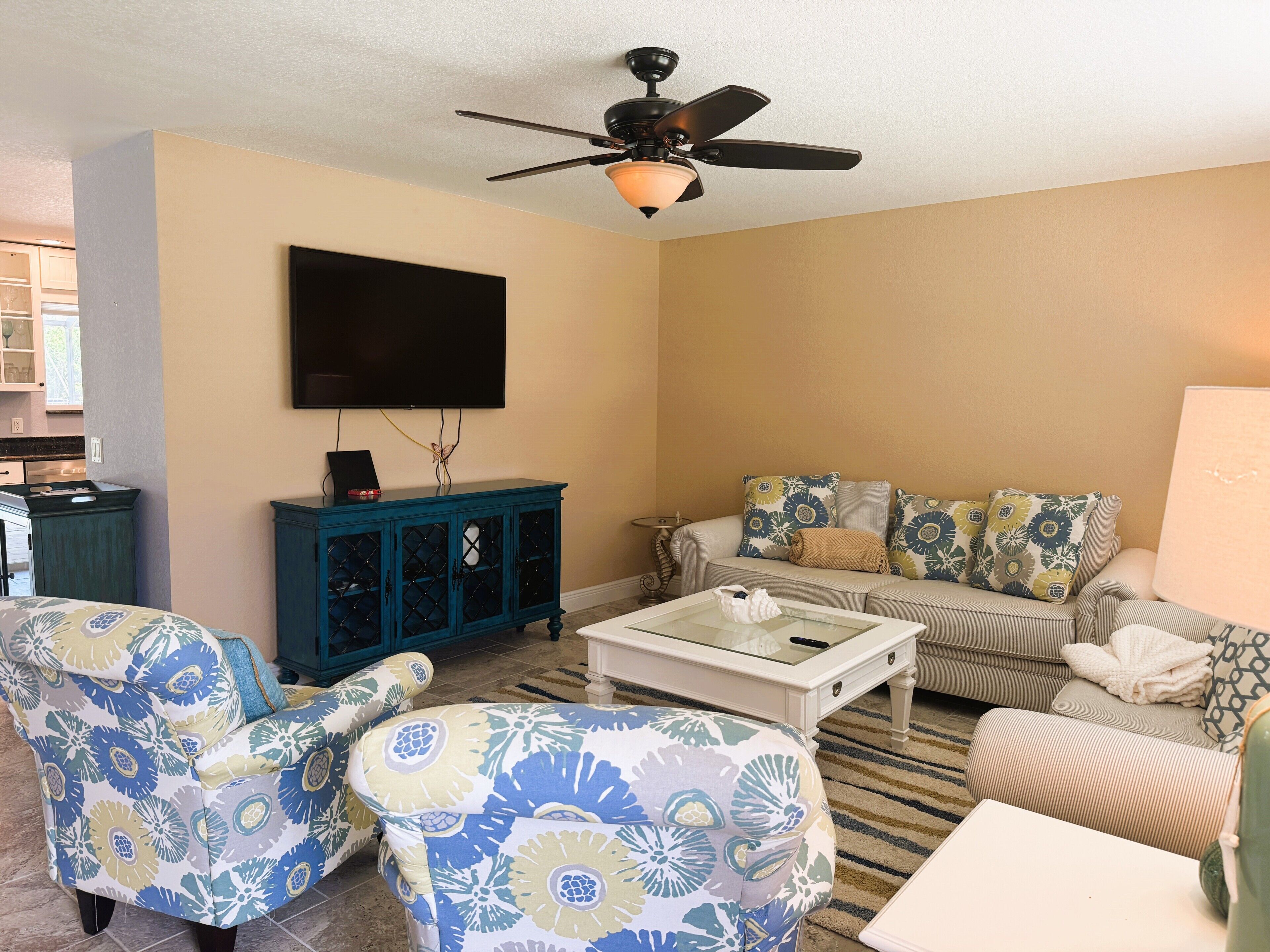 Ein Wohnzimmer mit Deckenventilator, einem flachbildfernseher an der Wand, einem blauen Schrank mit Glastüren, einem beige farbenen Sofa mit gemusterten Kissen und einem weißen Couchtisch.