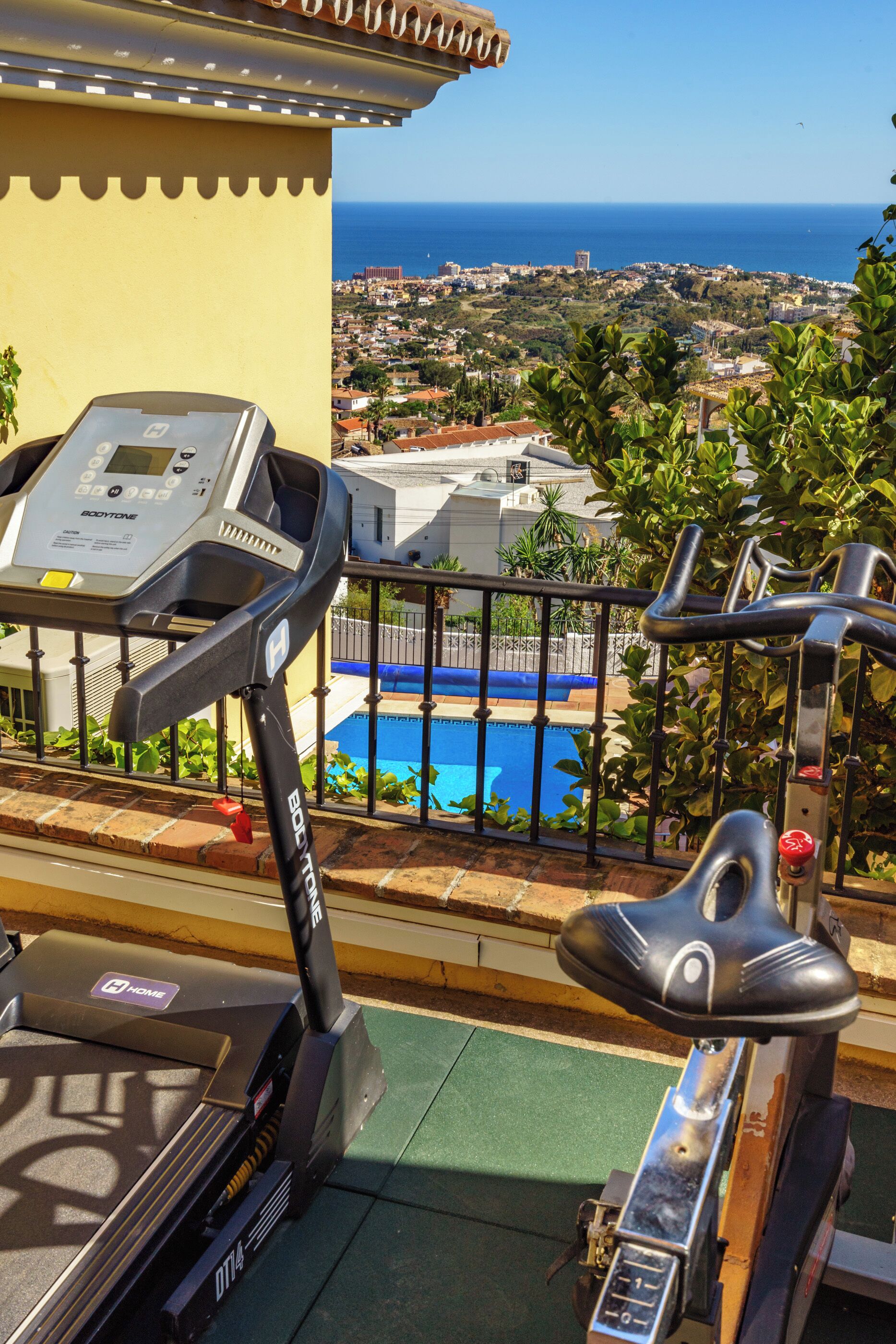 Un vélo d’appartement, doté d’un écran numérique, ainsi qu’une piscine et une vue sur une ville côtière.