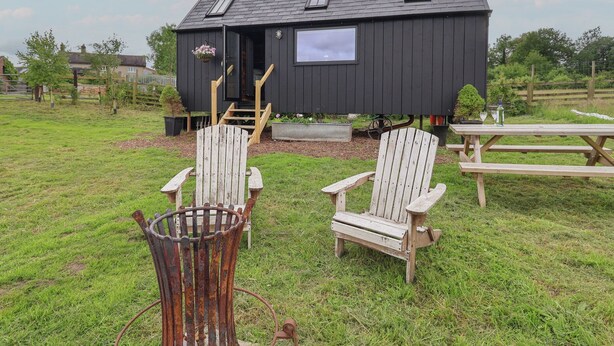 A small black cabin with a porch, two Adirondack chairs, a picnic table, and a metal fire pit.