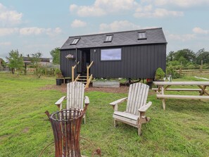 Eine kleine schwarze Holzhütte mit Veranda, zwei Adirondack-Stühlen, einem Picknicktisch und einem Metall-Feuerkorb.