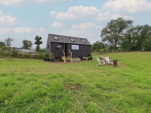 En lille, mørk træhytte med skråt tag, en trætrappe og et bord med stole i en græsmark.