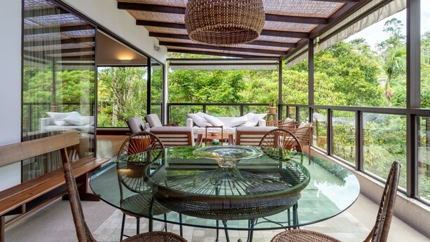 A modern outdoor dining area with a glass table, wicker chairs, and a hanging light fixture. - Casa com Lareira e Piscina Perto da Granja Comary (Teresópolis)