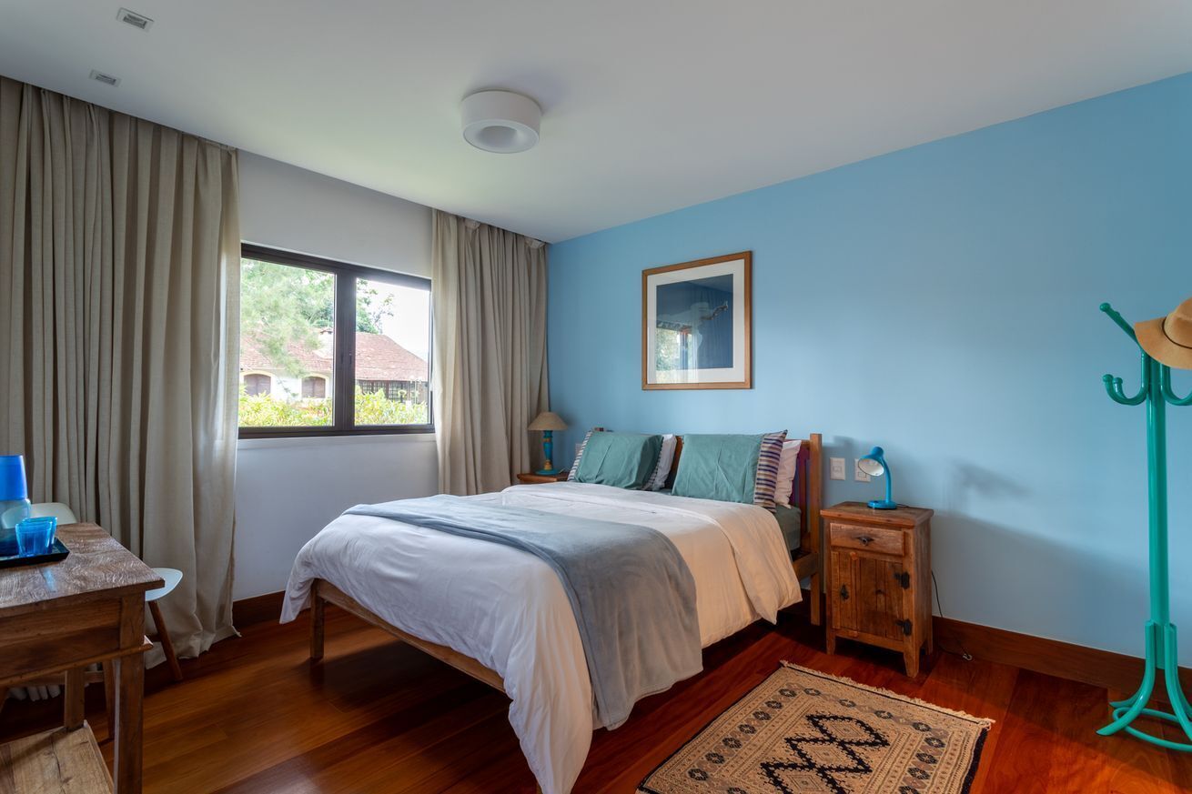 Quarto com cama, mesa de cabeceira, luminária, janela com cortinas, piso de madeira e parede azul.