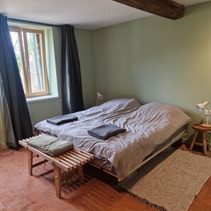 Ein Schlafzimmer mit Bett, einer Bank, einem kleinen Hocker, einem Fenster mit Vorhängen und einer Lampe.
