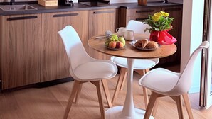 Una cocina moderna con armarios de madera, una cafetera y un comedor con sillas blancas y una mesa con frutas y pasteles.