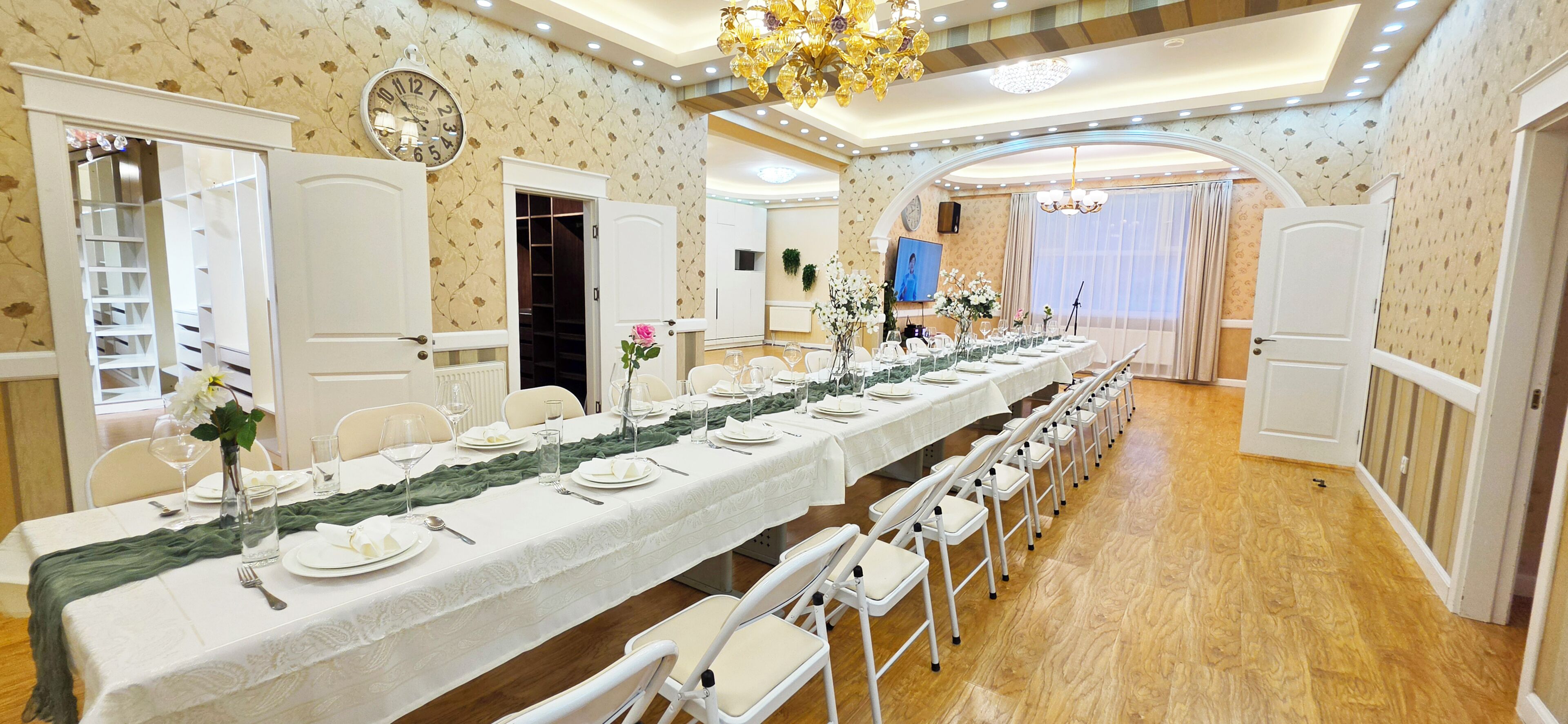 A long dining table with chairs, a chandelier, and a clock on the wall.