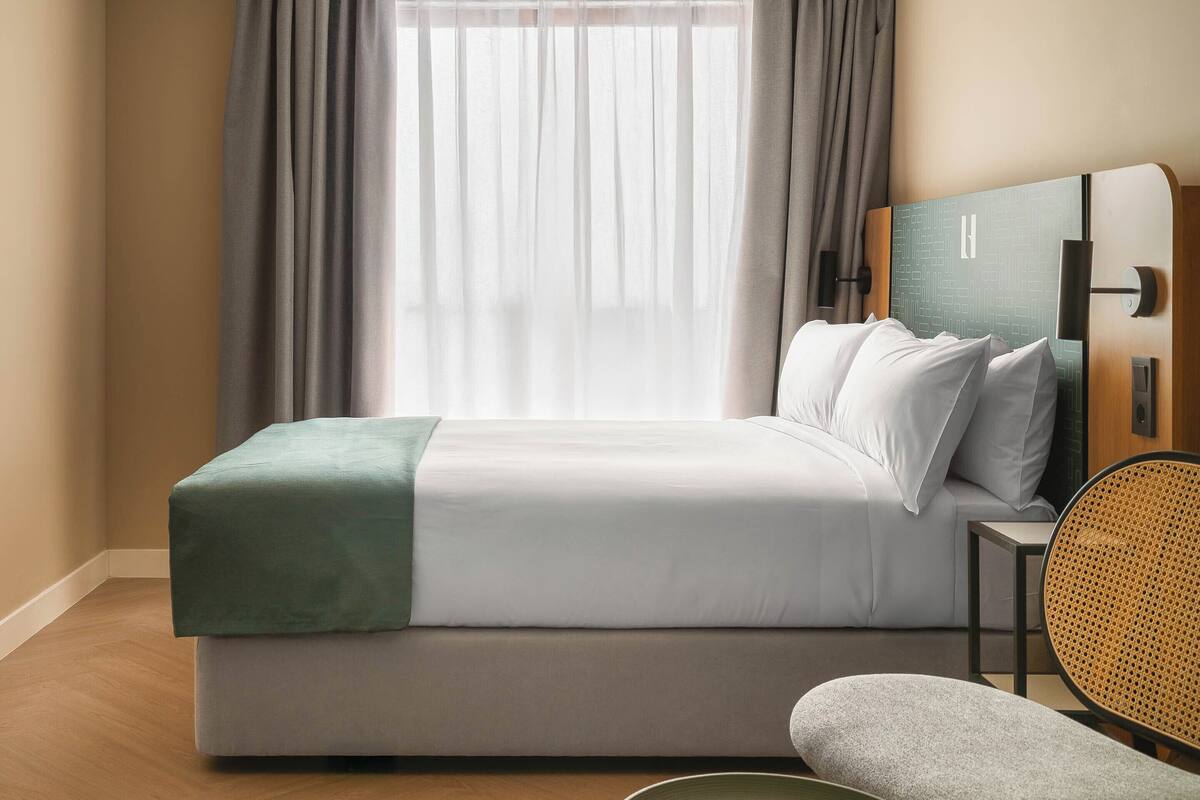 Ein modernes Hotelzimmer mit Bett, Nachttisch, Stuhl und einem Fenster mit Vorhängen.