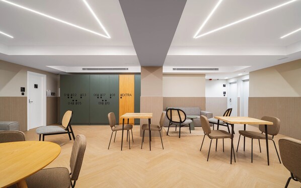 A modern conference room with a herringbone floor, wooden tables, and chairs, a herringbone-patterned wall, and a door with numbered lockers. - Limehome Madrid Ventas (Madrid)