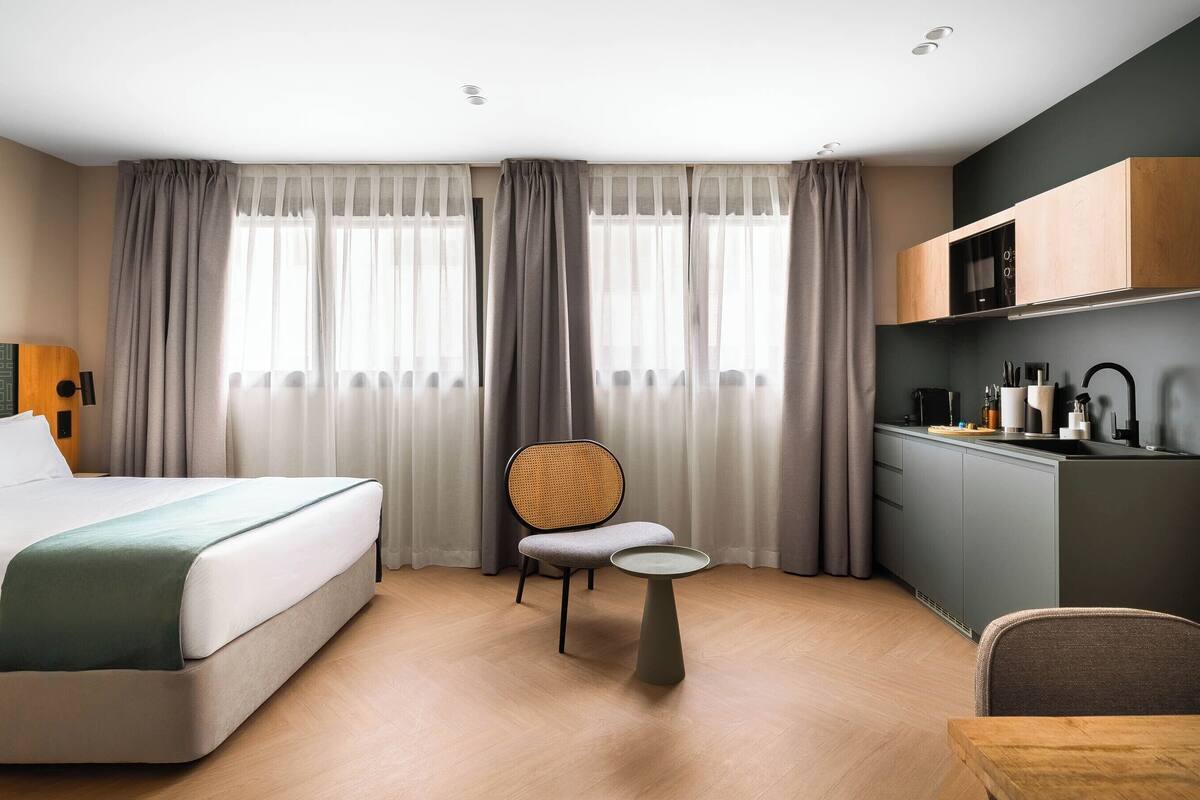Ein Hotelzimmer mit Bett, kleinem runden Tisch, Stuhl, Küchenzeile und einem Fenster mit Vorhängen.