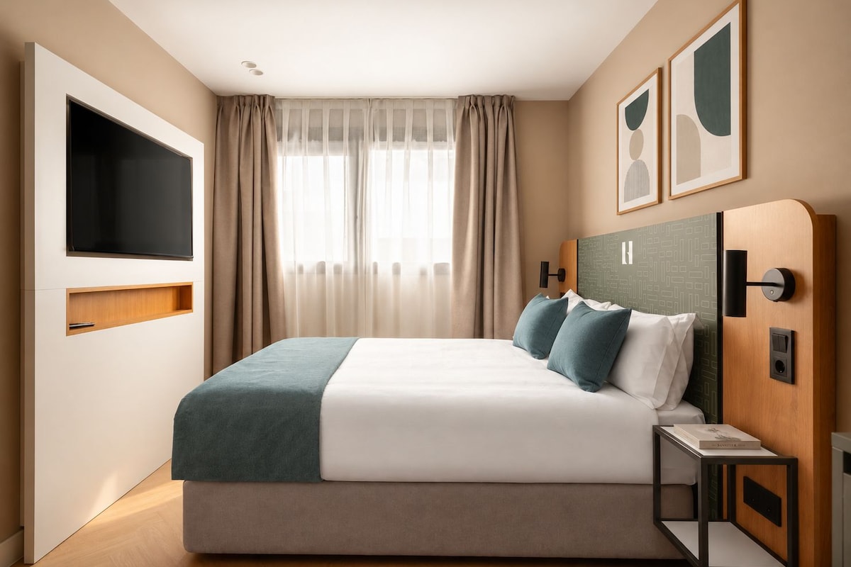 Ein modernes Hotelzimmer mit einem großen Bett, einem Flachbildfernseher, einem Nachttisch und einem Kopfteil mit integrierten Bedienelementen.