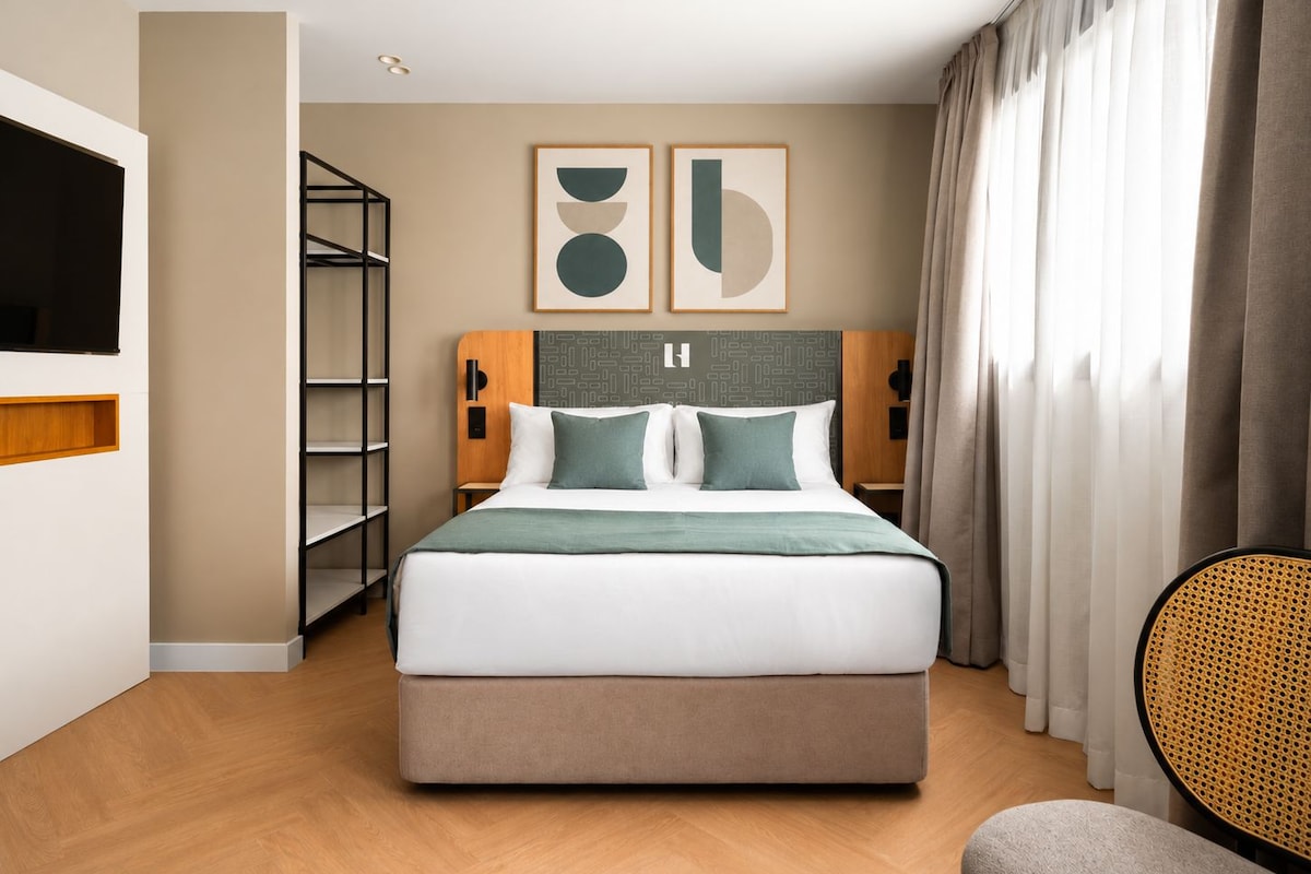 Ein modernes Hotelzimmer mit einem großen Bett, einem Fernseher, einem Bücherregal und einem Fenster mit Vorhängen.