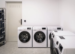 Un lavadero con tres lavadoras y secadoras de carga frontal, un tendedero de metal con ropa doblada y una puerta con cerradura electrónica.