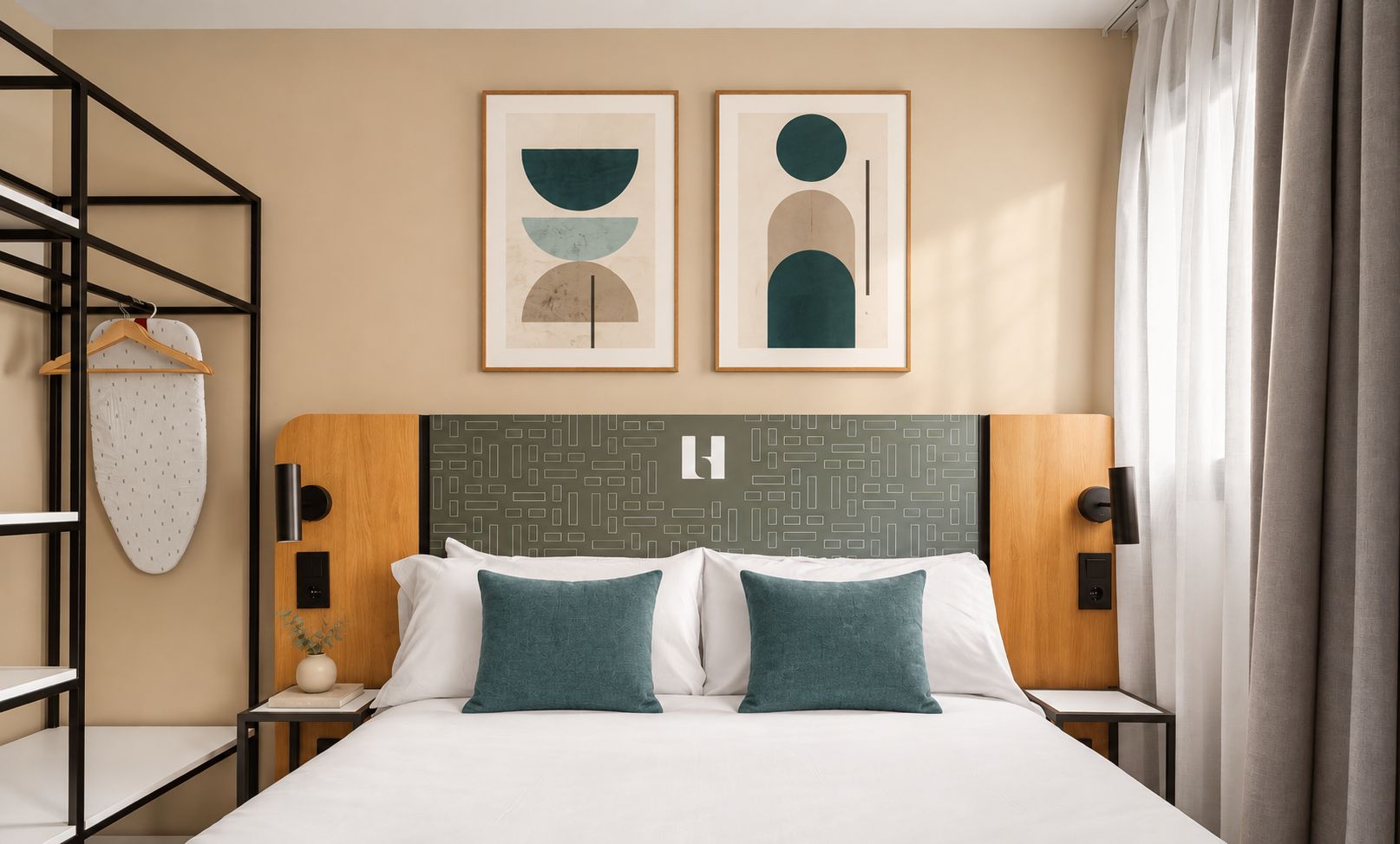 Habitación de hotel con cama, dos almohadas, cabecero con diseño geométrico, estante con una prenda colgada y dos obras de arte abstractas enmarcadas.