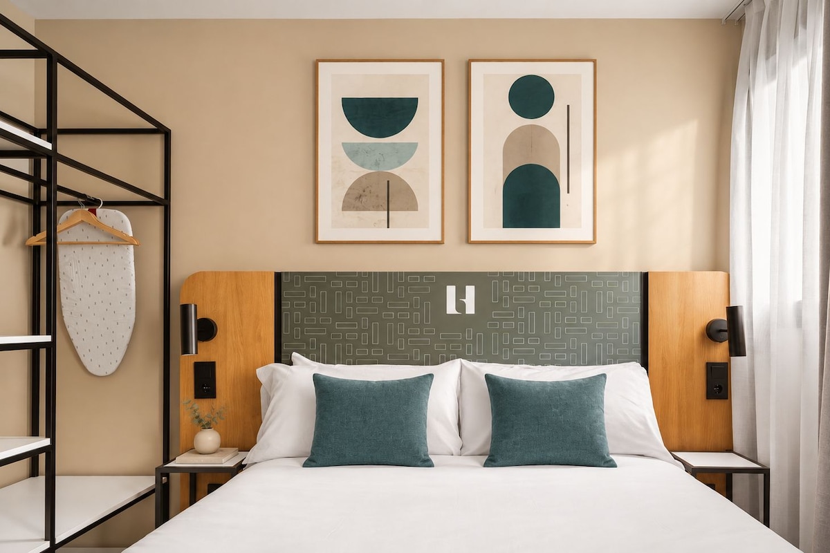 Ein Hotelzimmer mit einem Bett, zwei Kissen, einem Kopfende mit geometrischem Muster, einem Wandregal mit einem aufgehängten Kleidungsstück und zwei gerahmten abstrakten Kunstwerken.