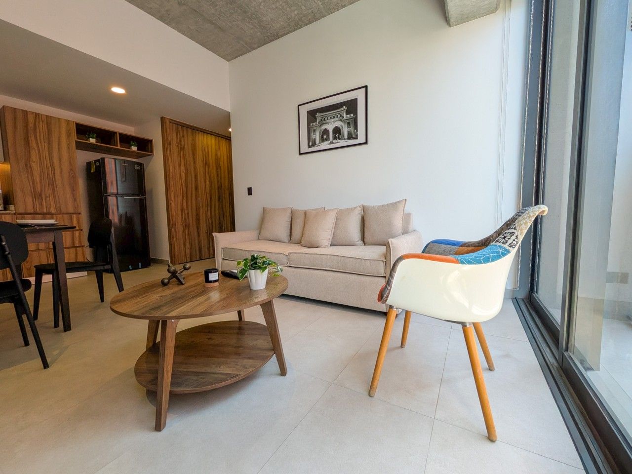 A modern living room with a sofa, a coffee table, and a chair. There is a dining area with chairs and a table. A refrigerator is visible in the background.