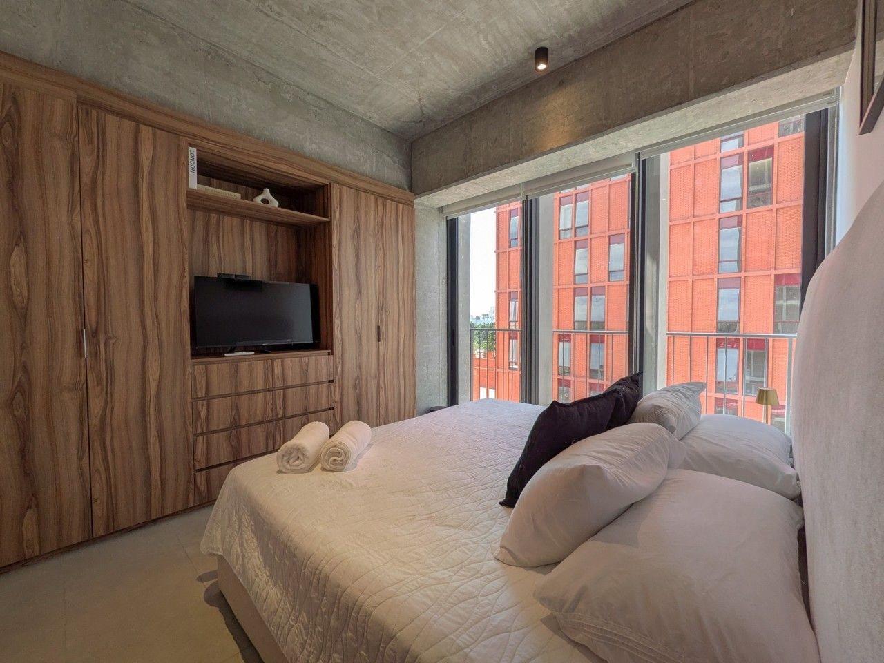 A modern hotel room with a large bed, a flat-screen TV, and a view of a red building through large windows.