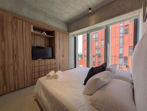 Ein modernes Hotelzimmer mit einem großen Bett, einem Flachbildfernseher und Blick auf ein rotes Gebäude durch große Fenster.