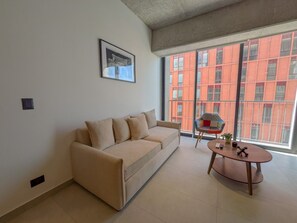 Een woonkamer met een beige bank, een klein rond tafeltje en een raam met uitzicht op een rood gebouw.