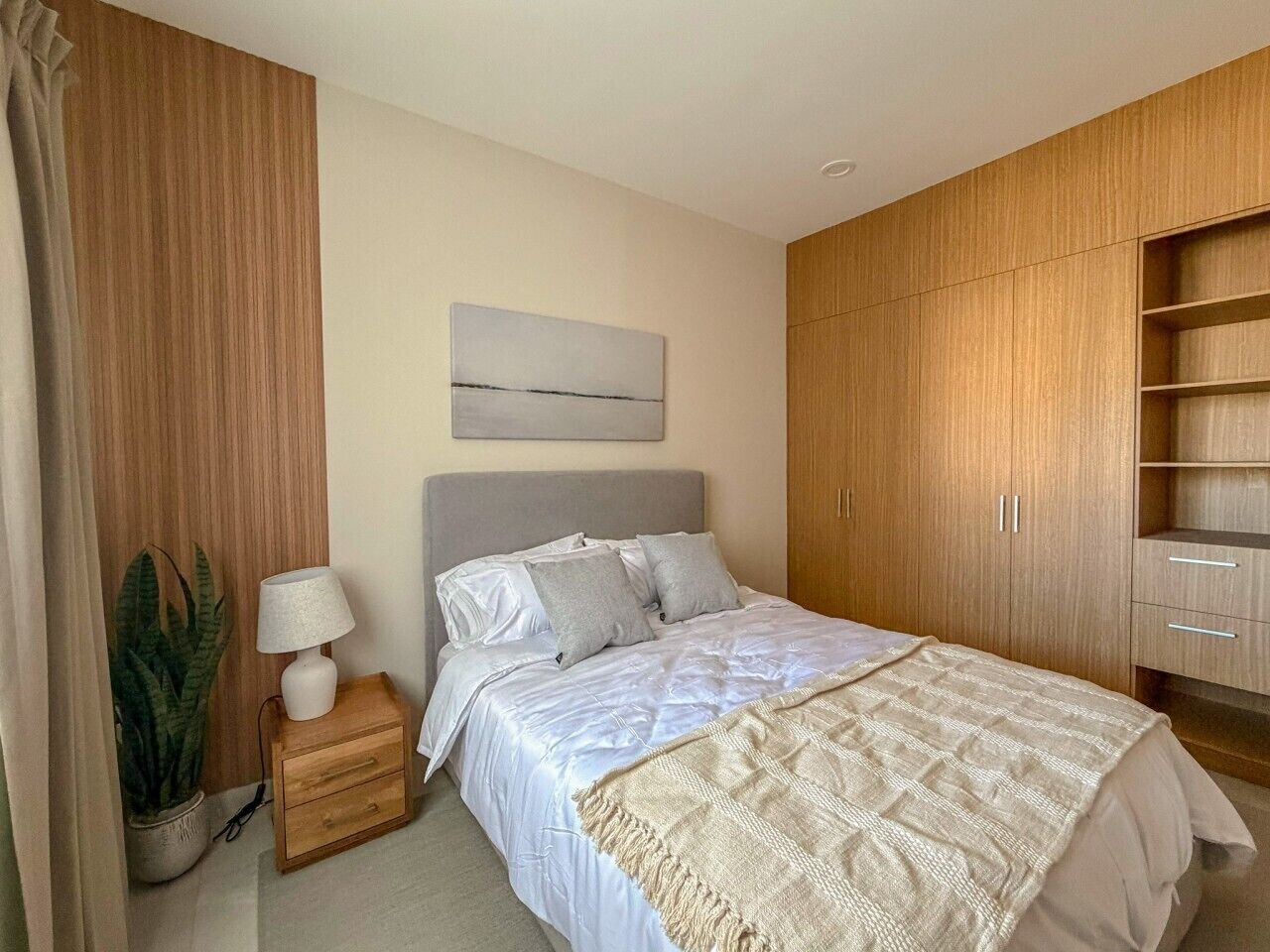 Un dormitorio con cama, mesita de noche, lámpara, planta en maceta y armario de madera.