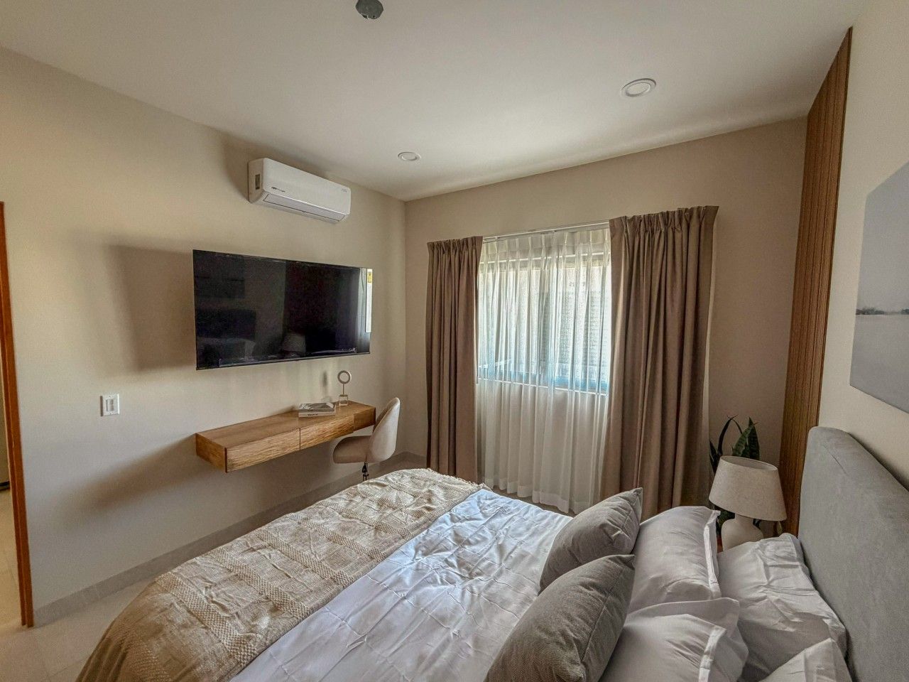 Une chambre d’hôtel avec un lit, un téléviseur fixé au mur, un bureau et une fenêtre avec des rideaux.