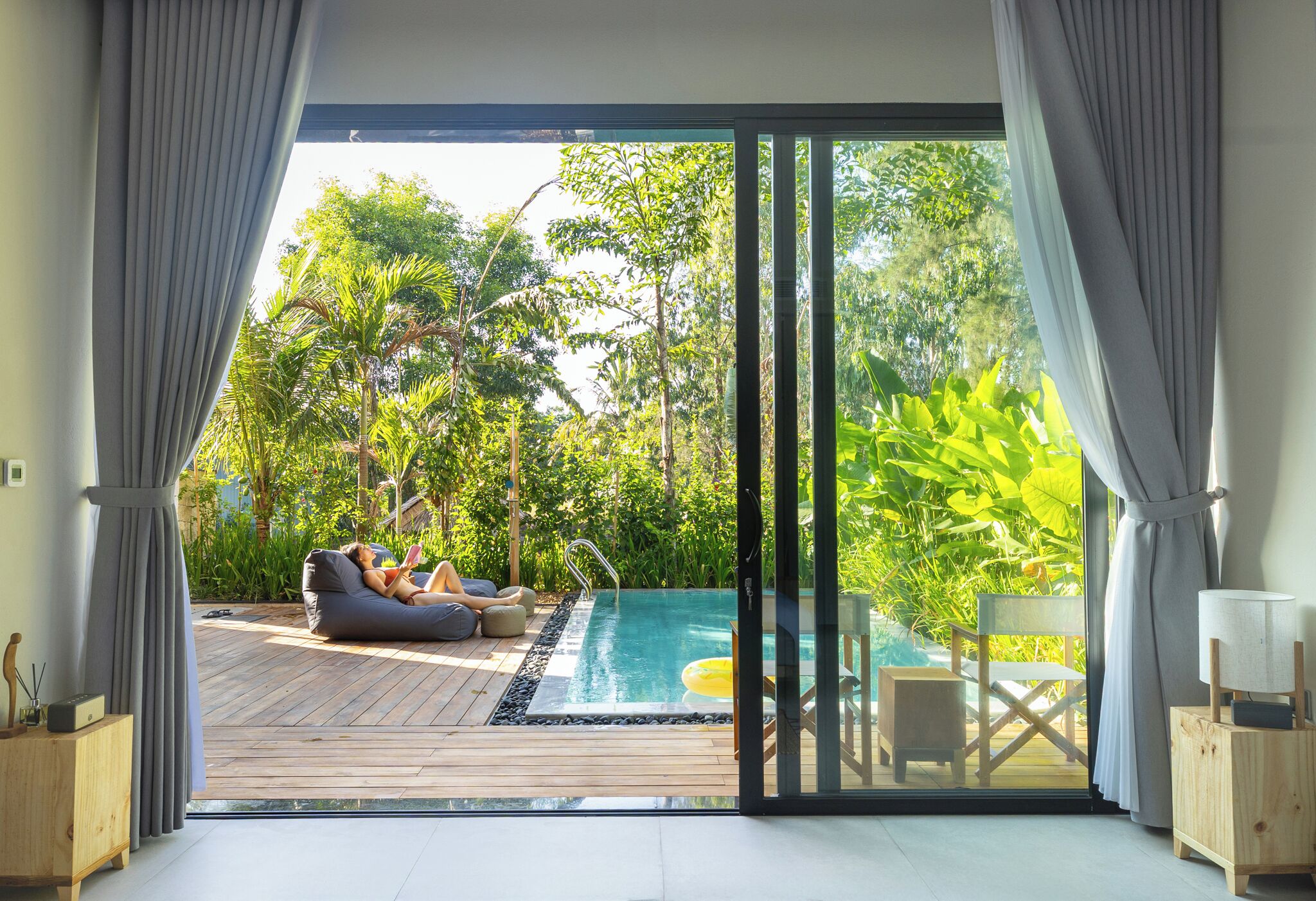Ein modernes Zimmer mit einer großen Glasschiebetür, die zu einem Außenpoolbereich mit Holzterrasse, Sitzgelegenheiten und üppiger Bepflanzung führt.