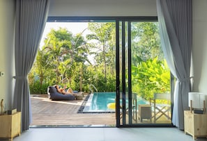 Ein modernes Zimmer mit einer großen Glasschiebetür, die zu einem Außenpoolbereich mit Holzterrasse, Sitzgelegenheiten und üppiger Bepflanzung führt.