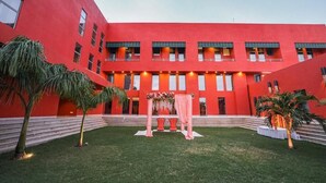 A red building with multiple windows and balconies, a central archway with floral decorations, and a small lawn area. - Gulab Kothi by Grapevine Varanasi (Varanasi)