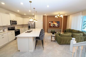 A modern kitchen with a central island, a dining area with a sofa, and a living room with a fireplace. - Modern 5 Bedroom Townhome at Windsor Cay 89BL (Clermont)