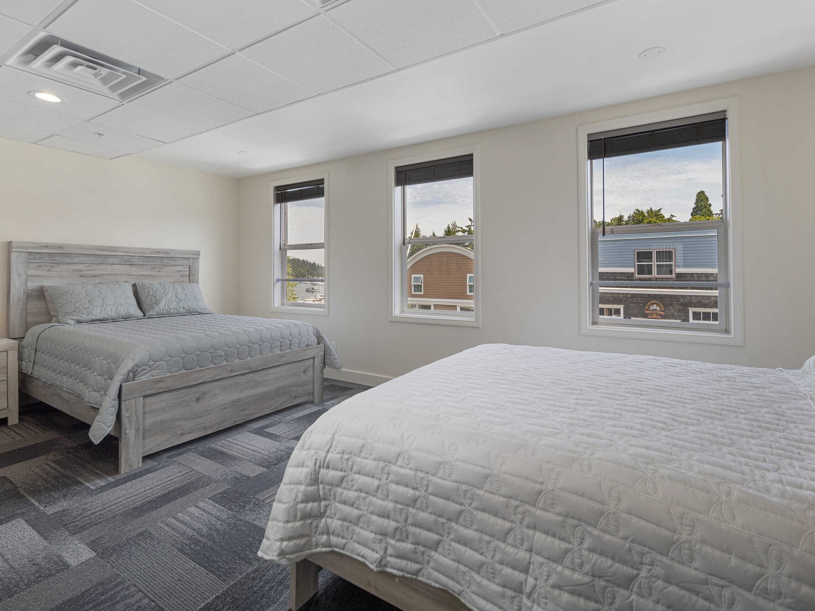 A bedroom with two beds, a wooden headboard, and large windows with views of buildings and trees.