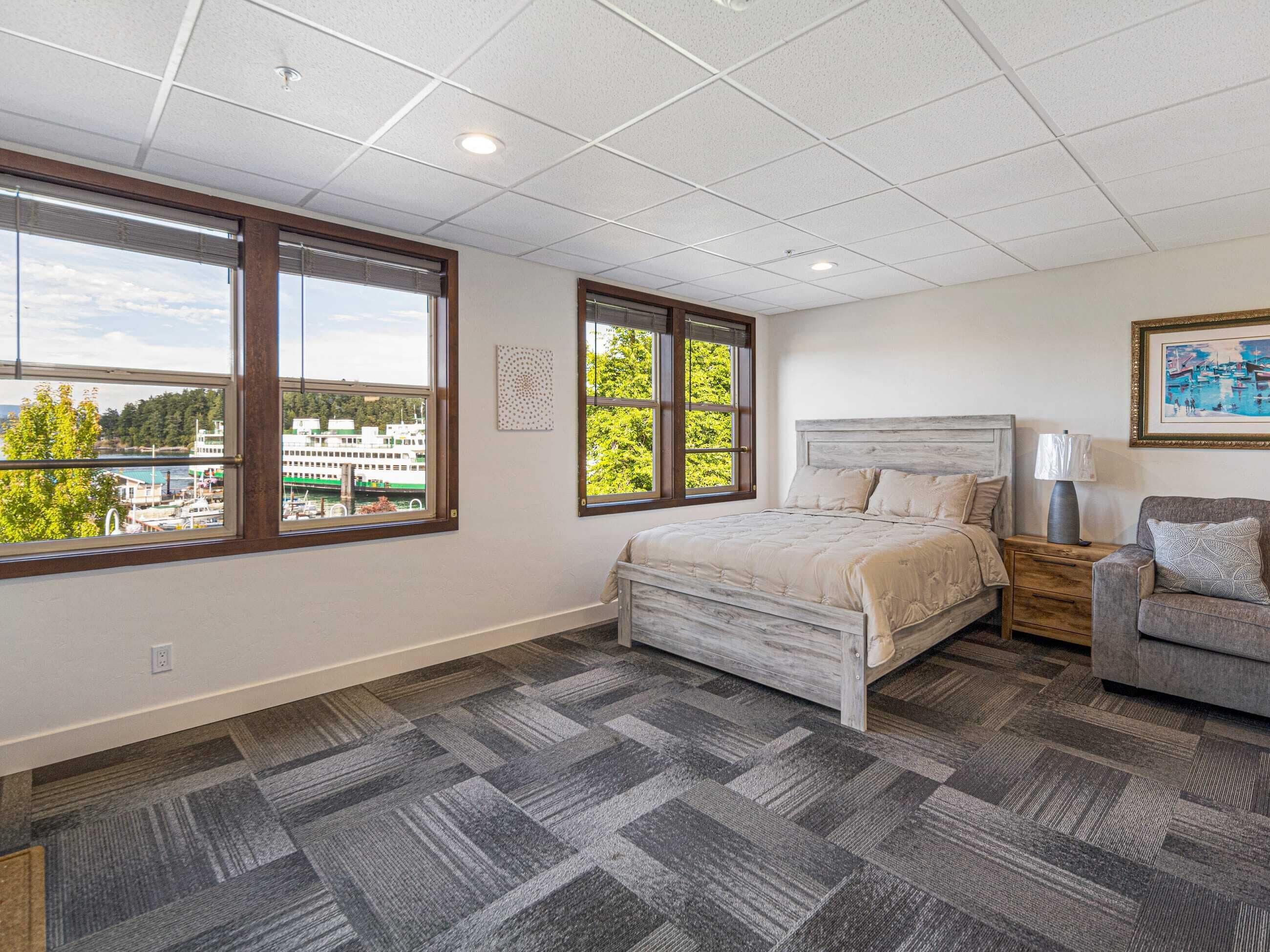A bedroom with a bed, a nightstand, a lamp, a chair, and large windows with a view of buildings and trees.