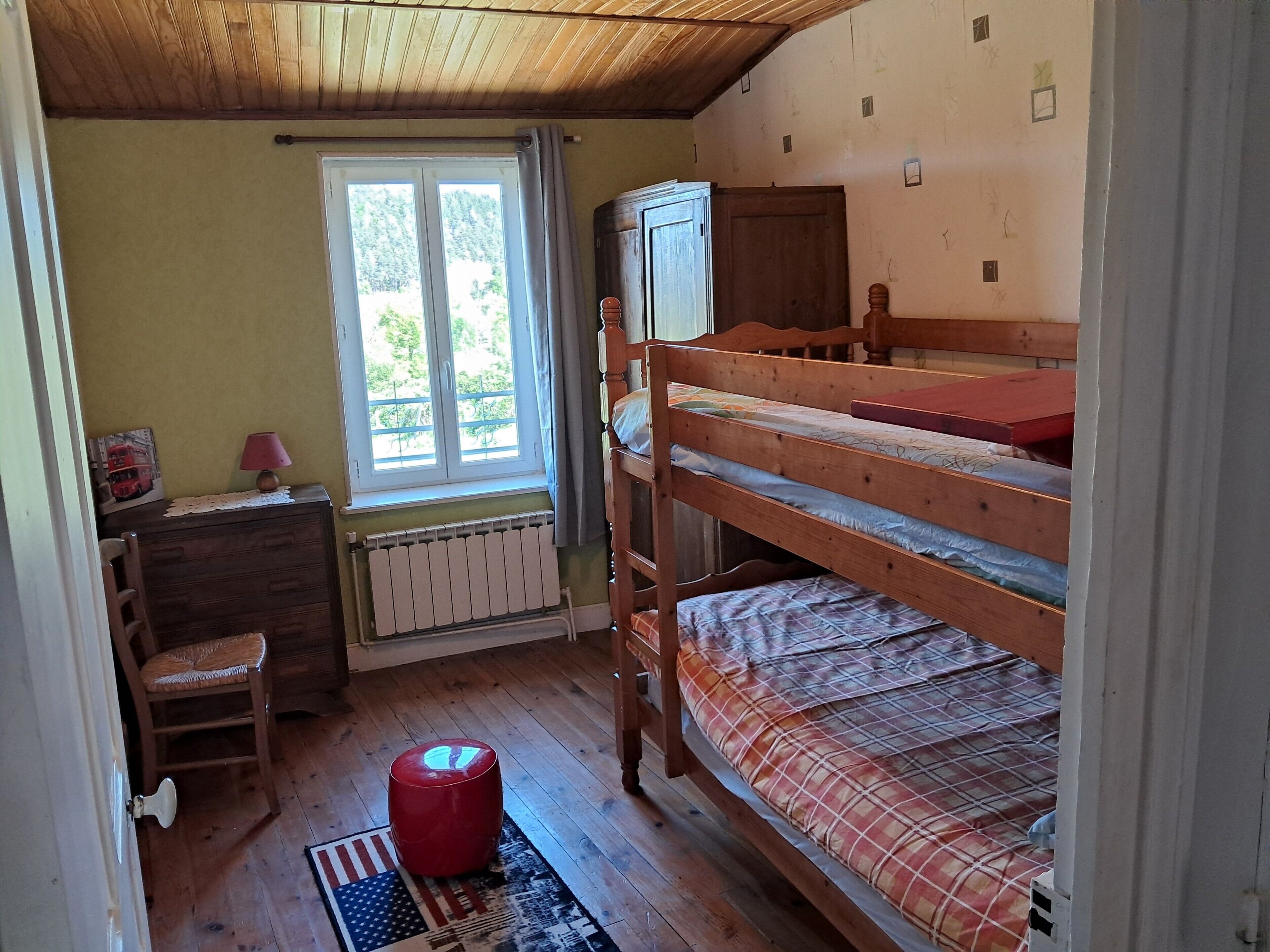 A room with a bunk bed, a wooden desk, a chair, a radiator, and a window with a view of greenery.