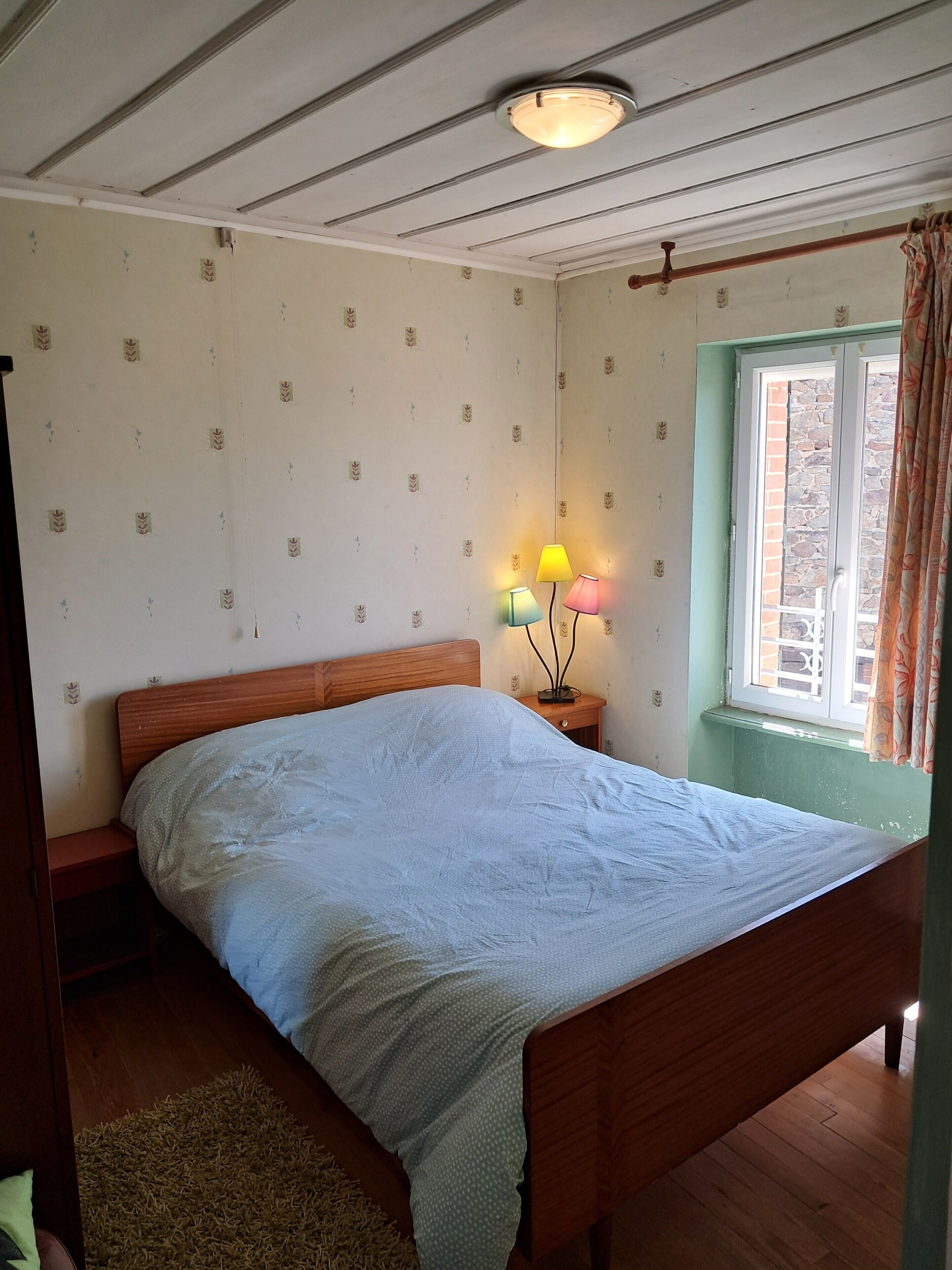 Une chambre à coucher comprenant un lit, une table de chevet, une lampe, une fenêtre avec des rideaux et un luminaire au plafond.