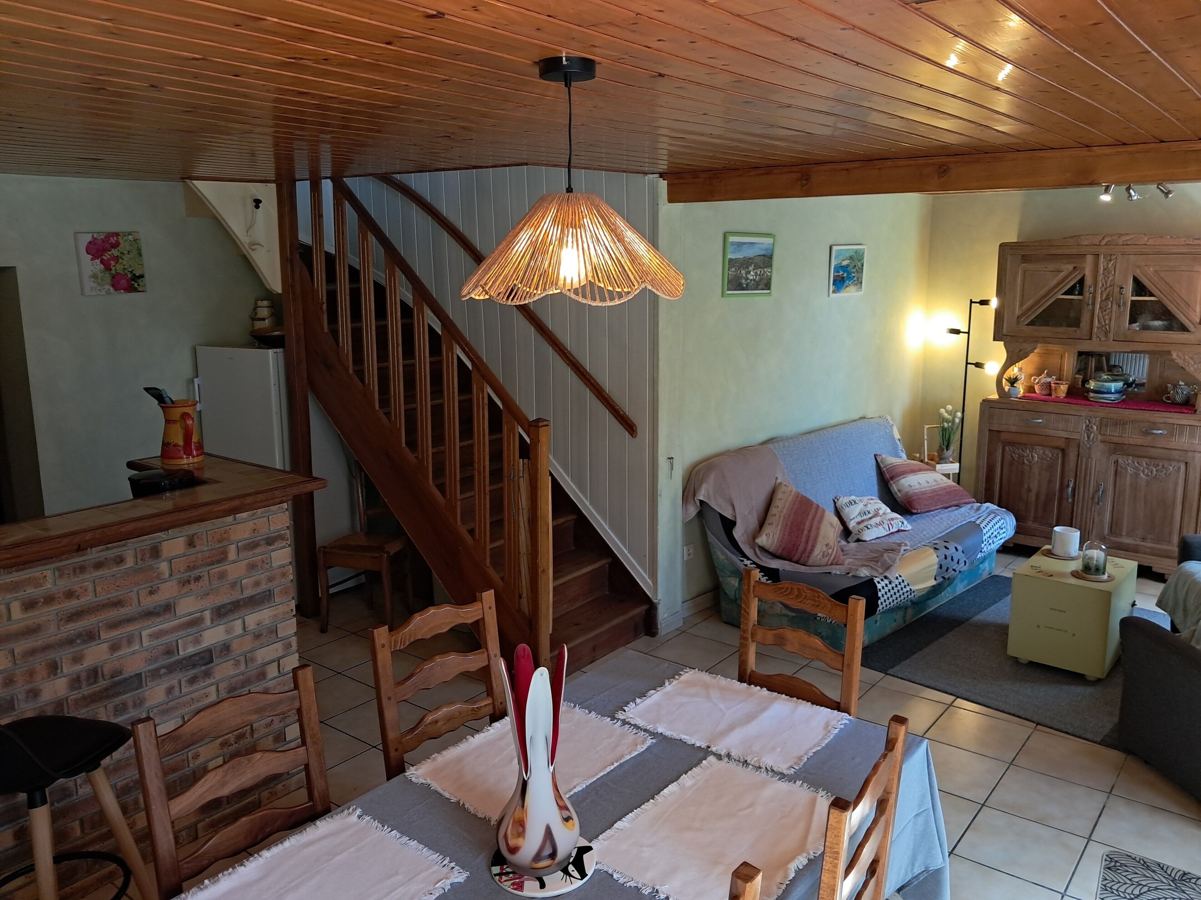 A cozy living room with a wooden staircase, a dining area with a table and chairs, a sofa, and a kitchen area with cabinets and a countertop.