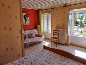 A bedroom with a wooden bed, a colorful sofa, a chair, a radiator, and a window with a view of the outdoors. - Holiday Home 'Gîte Les Glycines' (Auzon)