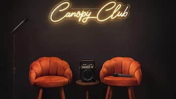 Two velvet armchairs facing each other with a vintage radio and microphone on a small table between them, against a dark background with a neon sign reading 'Canopy Club'.