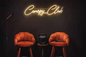 Two velvet armchairs facing each other with a vintage radio and microphone on a small table between them, against a dark background with a neon sign reading 'Canopy Club'.