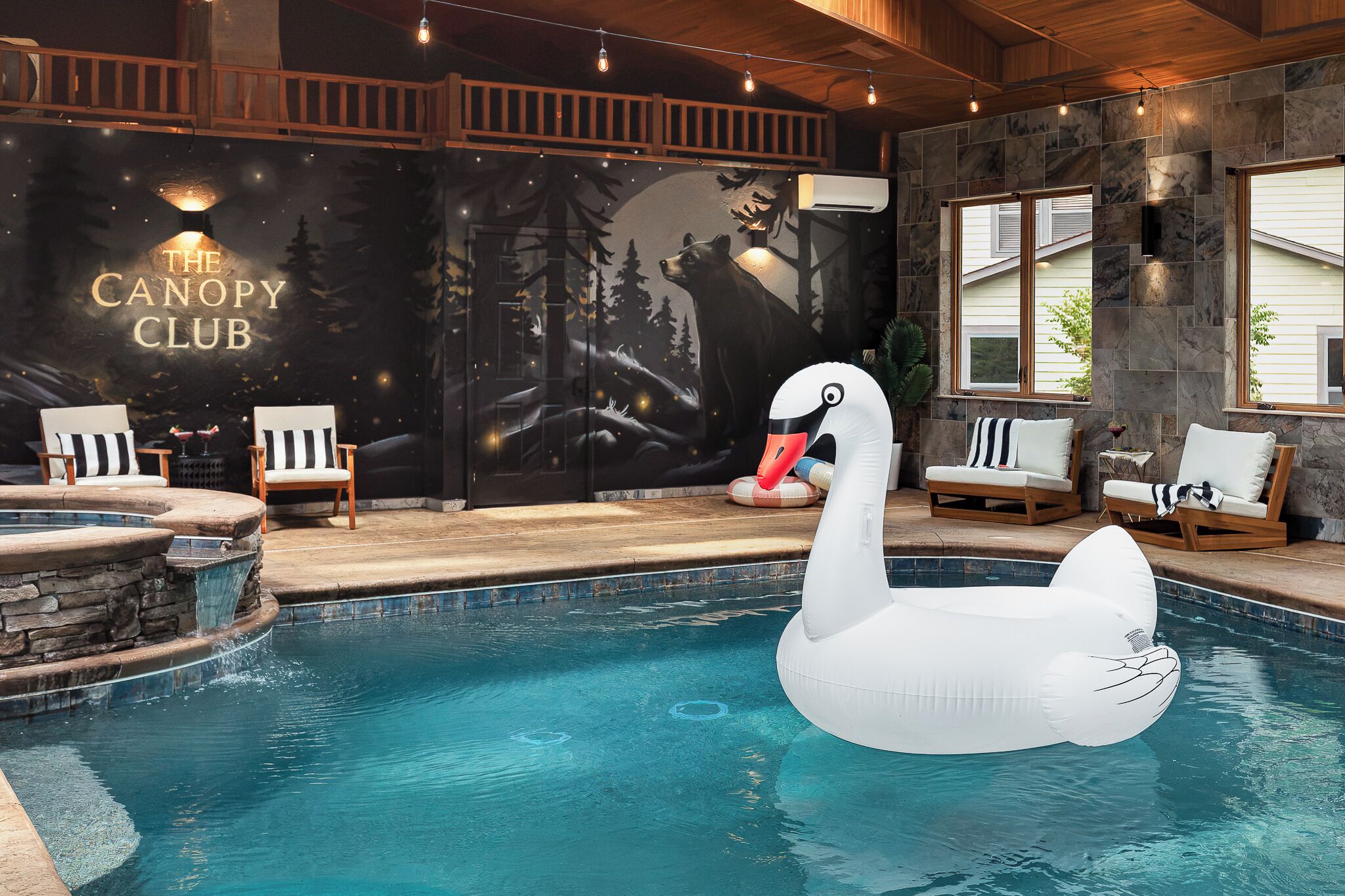 Une piscine intérieure avec un flotteur en forme de cygne, des chaises longues en bois et une fresque murale portant l’inscription « THE CANOPY CLUB ».