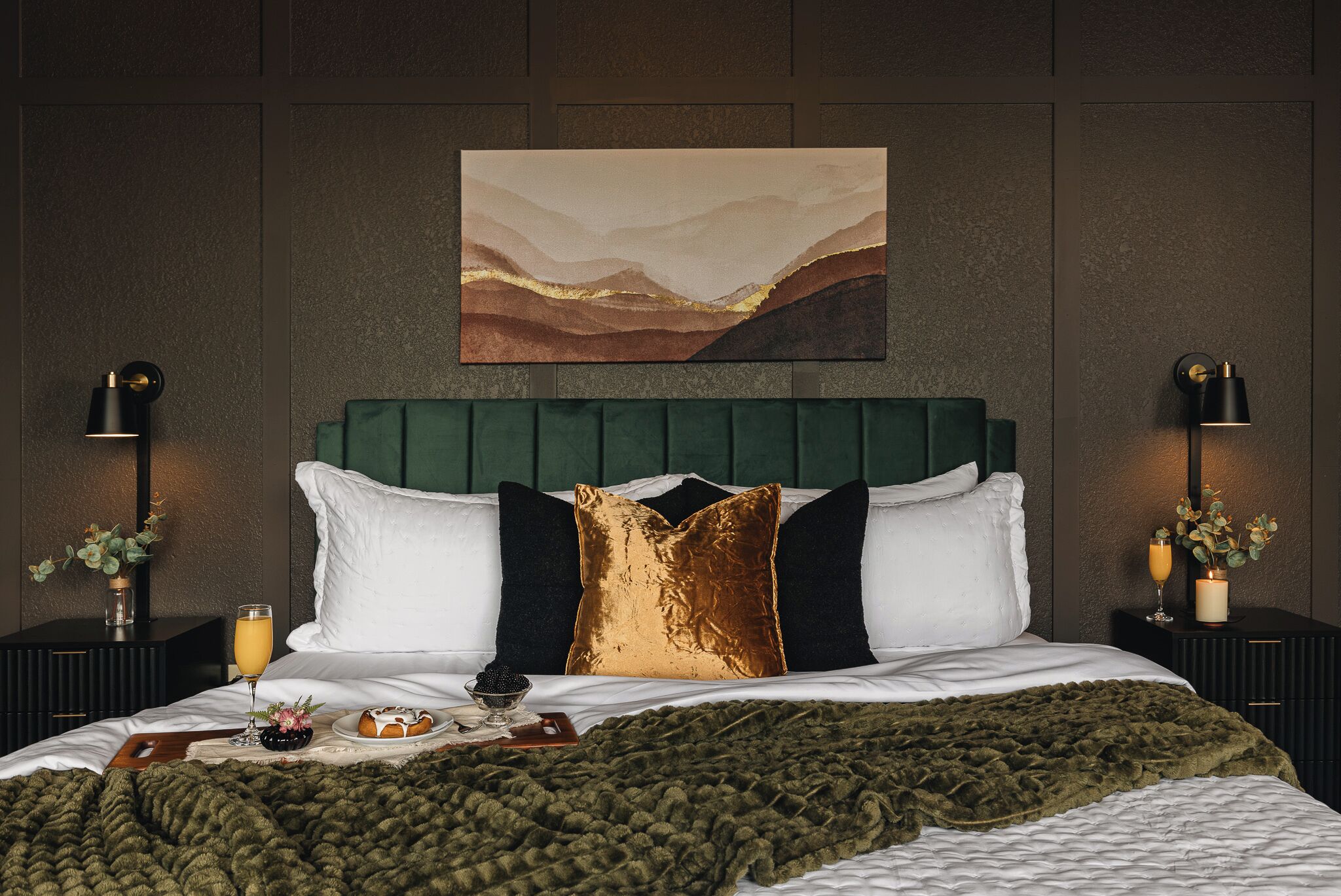 Ein Schlafzimmer mit einem grünen, mit Stoff bezogenen Kopfteil, weißen und goldenen Kissen, einer grünen Decke und einem Gemälde über dem Bett.