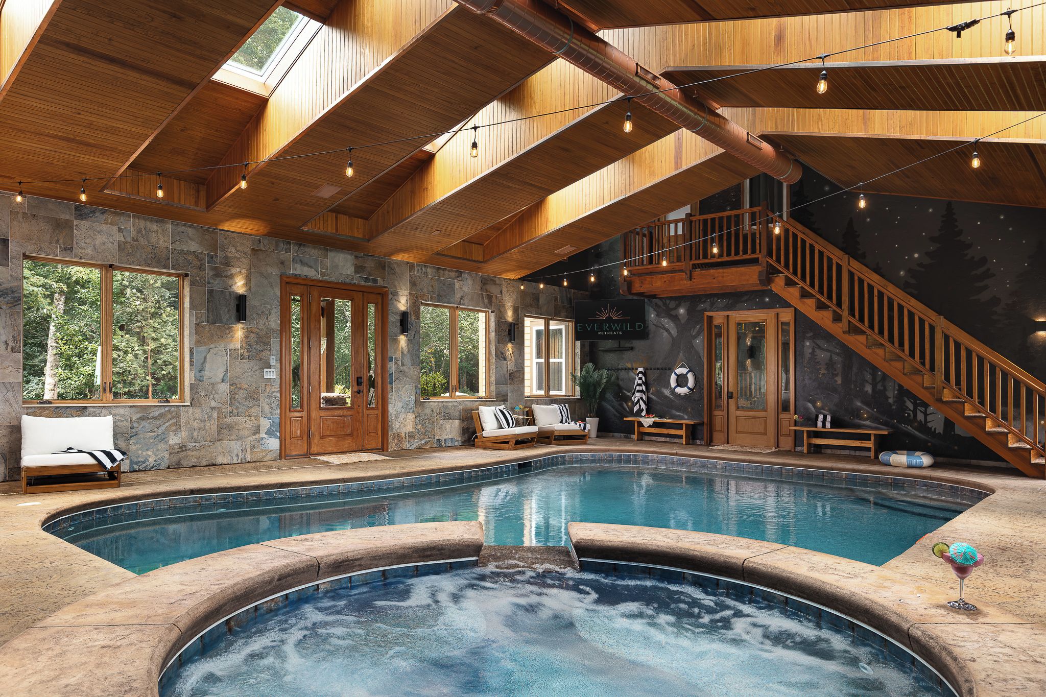 A large indoor swimming pool with a hot tub, wooden ceiling, stone walls, and a staircase leading to an upper level.