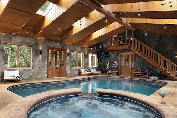 Una amplia piscina interior con un jacuzzi, techo de madera, paredes de piedra y una escalera que conduce a un nivel superior.