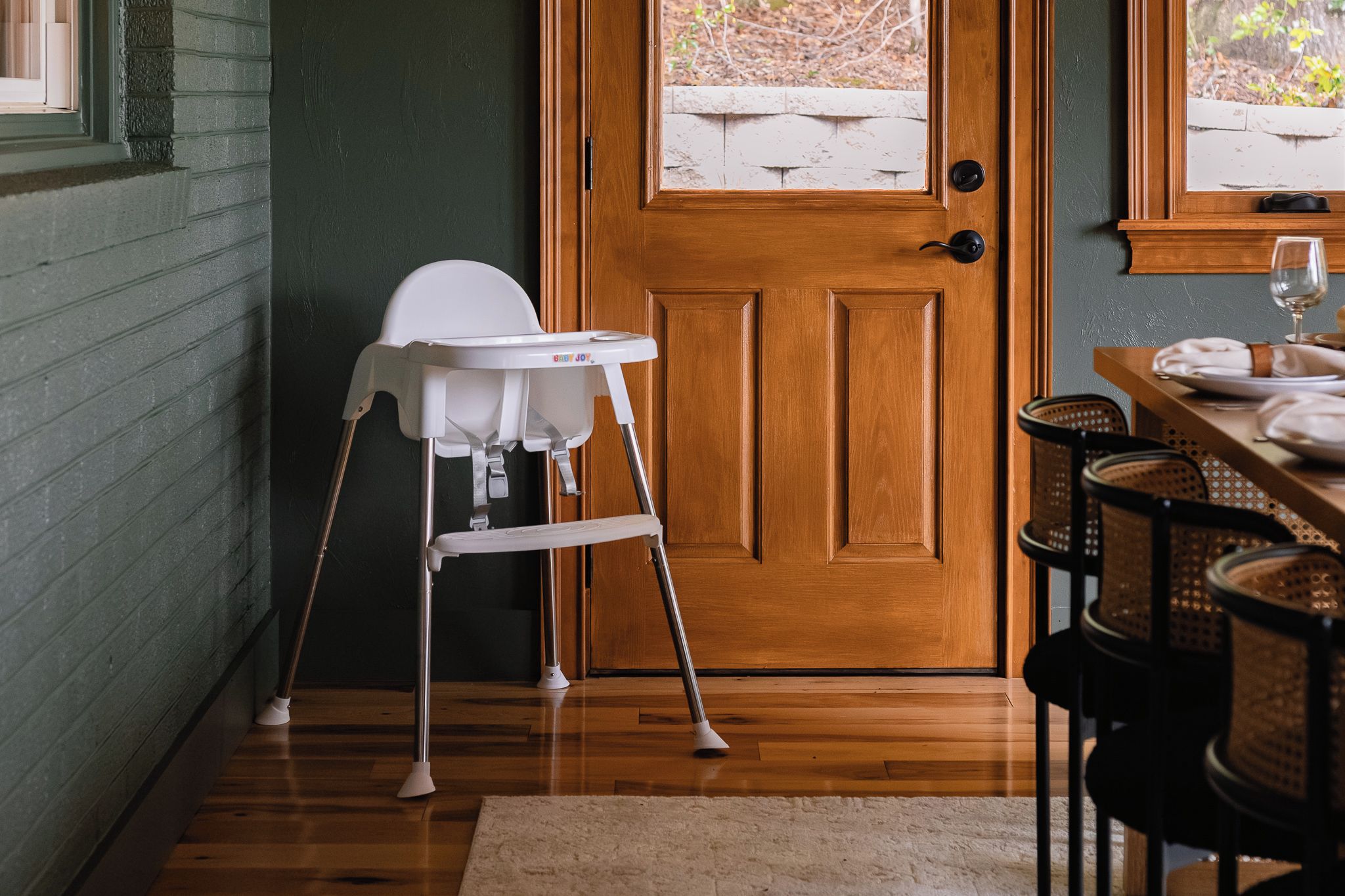 Una habitación con puerta de madera, una trona blanca y una mesa de comedor dispuesta con platos y una copa de vino.