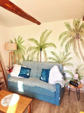 A room with a sofa, a lamp, a small table, and a wall with a palm tree mural. - Between the vineyards and the canal. Fully renovated 3-story house (Azille)