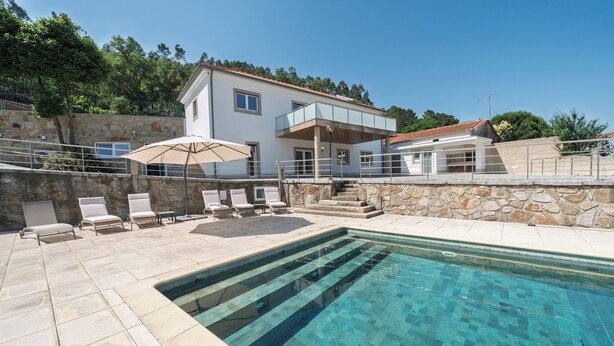 Un área junto a la piscina con una pileta cristalina, paredes de piedra y una casa de dos pisos con balcón.