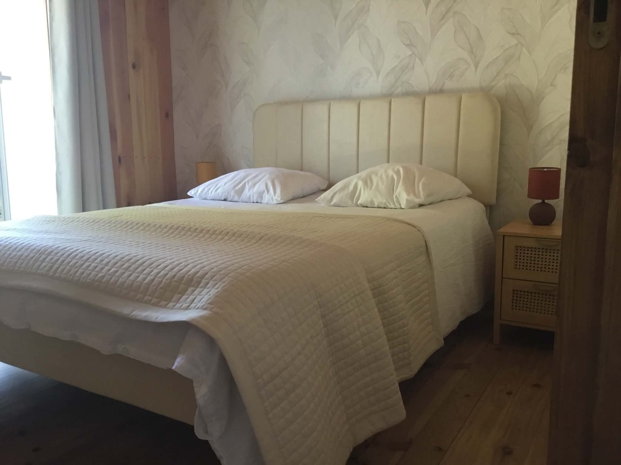 A bed with a white headboard and two pillows, a bedside table with a lamp, and a patterned wall.