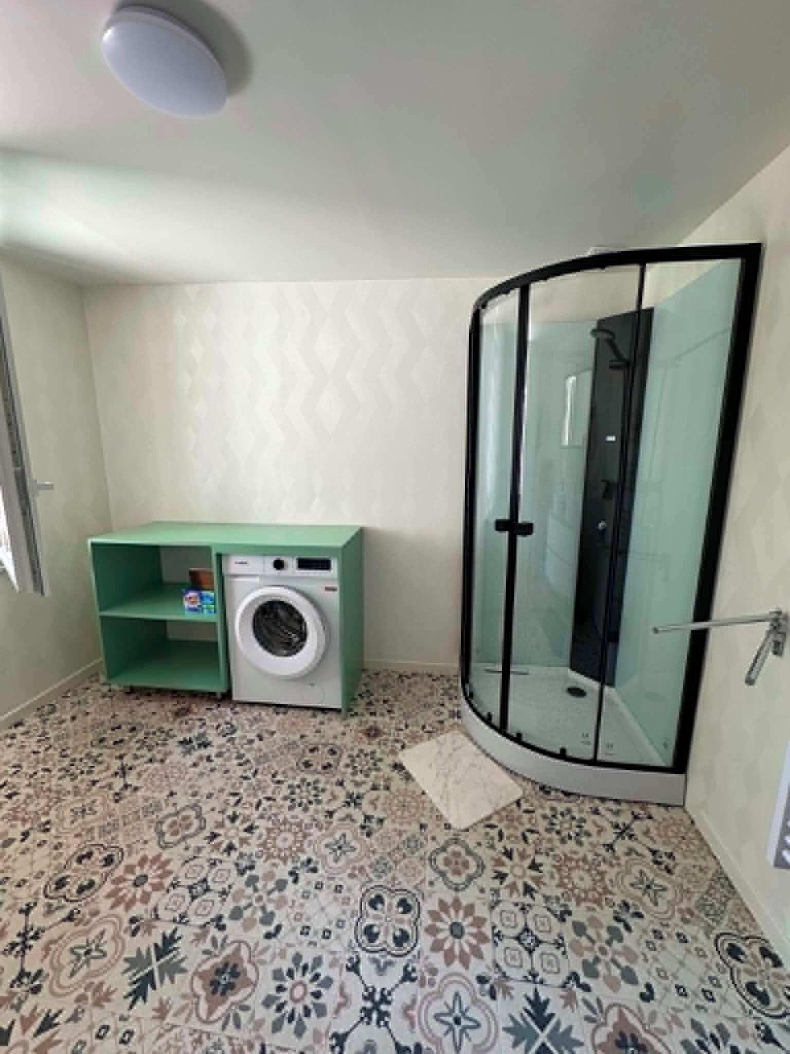Une petite salle de bain équipée d’un lave-linge, d’une cabine de douche en verre et de carreaux de sol à motifs.