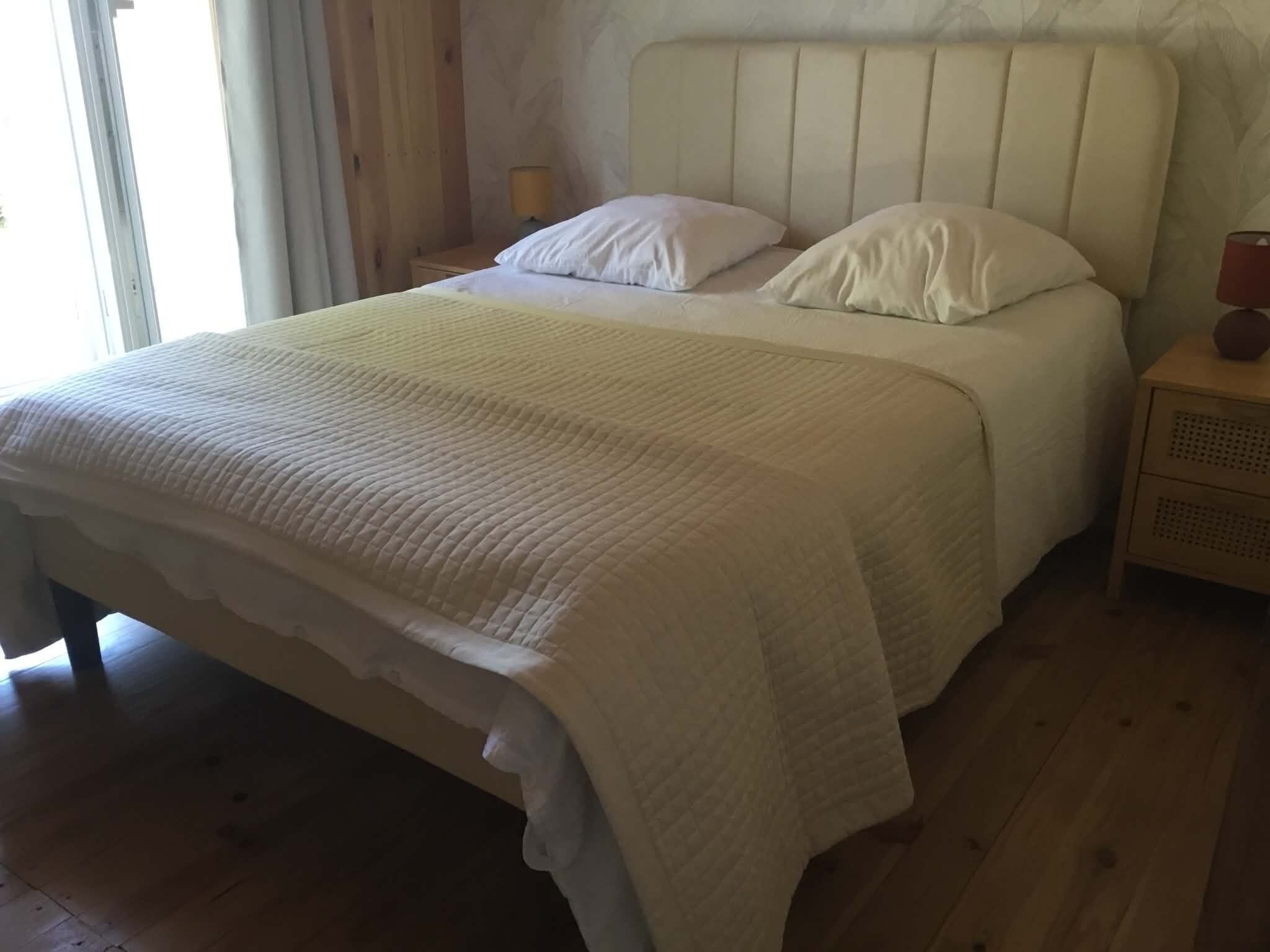 Una cama con una colcha blanca y almohadas, un cabecero de madera, una mesita de noche con una lámpara y una ventana con cortinas.