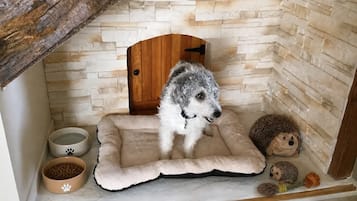 A cozy dog bed with a fluffy dog, a small wooden door, and two bowls with paw prints.