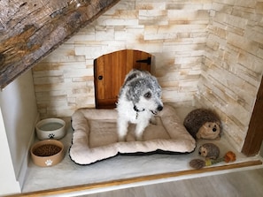 Una cama cómoda para perro con un perro esponjoso, una pequeña puerta de madera y dos comederos con huellas de pata.