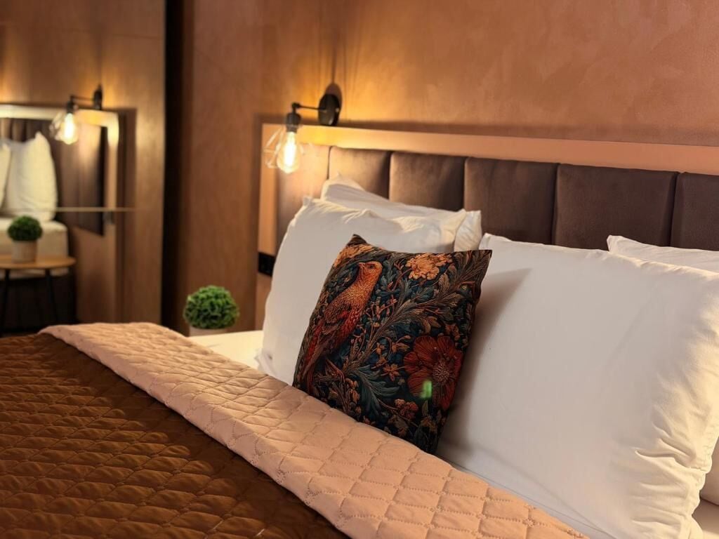 A neatly made bed with a quilted brown cover, a decorative pillow with a floral pattern, and a headboard with a light fixture.